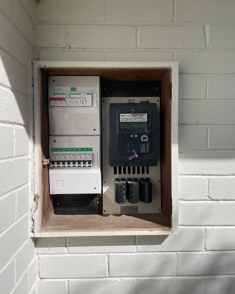 Electrical panel mounted on white brick wall containing circuit breakers, meters, and control switches in wooden enclosure