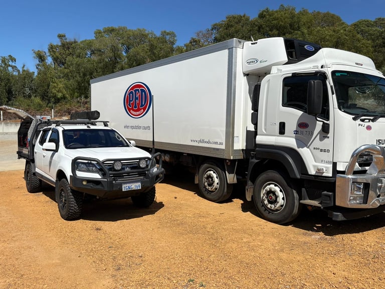White Isuzu truck with red logo and white vehicle parked on dirt ground with trees in background