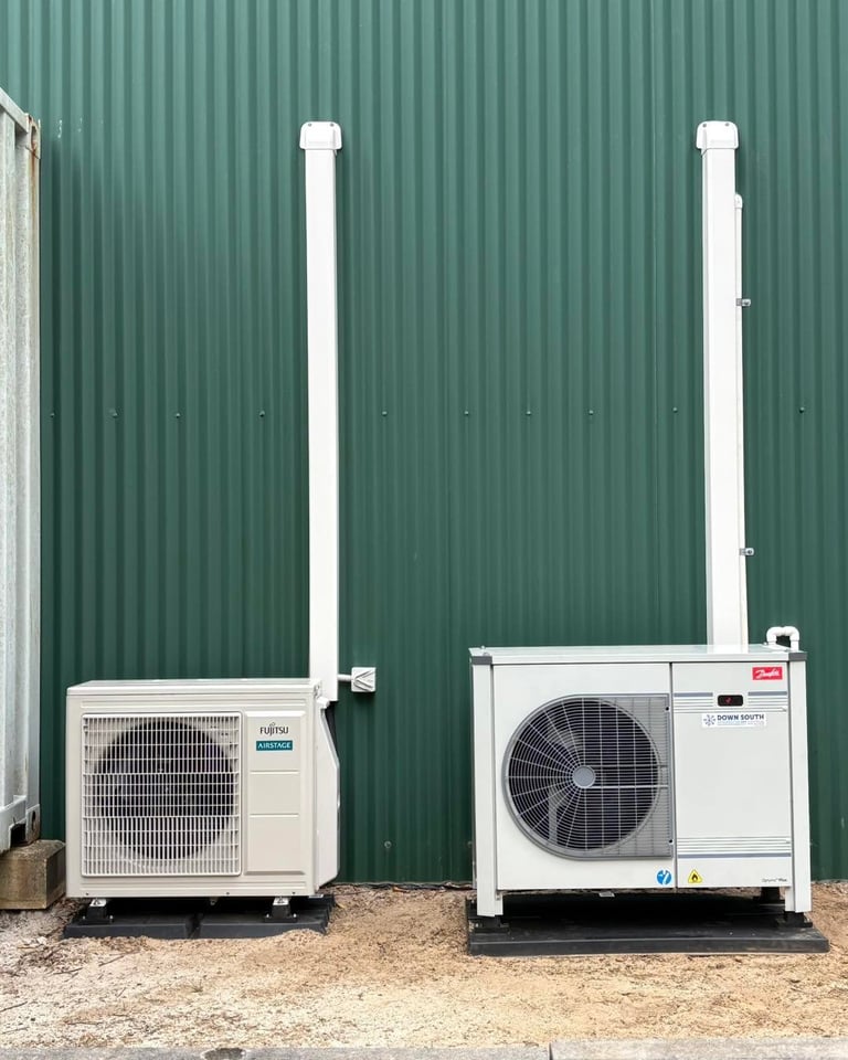 Two outdoor HVAC heat pump units against a green corrugated metal wall with white vertical pipes and condensers